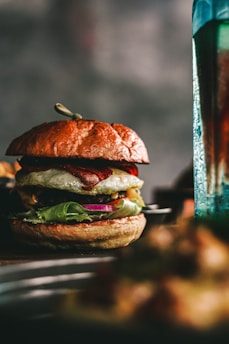 A close-up of a gourmet burger with a sesame seed bun, topped with lettuce, onion, tomato, cheese, a fried egg, and bacon. The background is slightly blurred, emphasizing the burger's rich textures and colors.