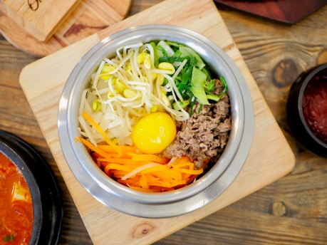 A Korean bibimbap dish is presented in a metal bowl on a wooden board. It contains colorful ingredients such as sliced carrots, bean sprouts, chopped greens, minced beef, and a raw egg yolk in the center. The table setting includes traditional Korean side dishes and a dish containing a red sauce.