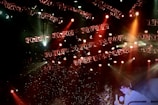 A stage lit with an array of bright spotlights and colored lights, featuring intricate light rigs and beams in red, orange, and white hues. The background is filled with sparkling effects, creating a lively and dynamic atmosphere. A person is partially visible, engaging in a performance or presentation.
