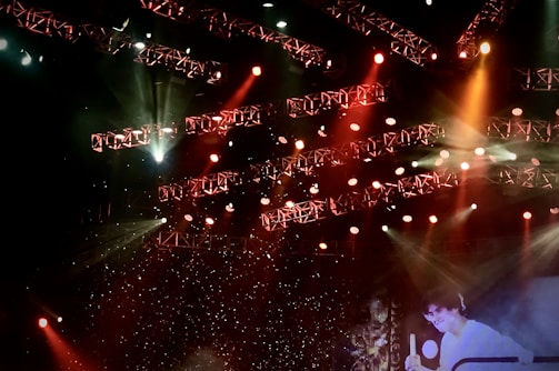 A stage lit with an array of bright spotlights and colored lights, featuring intricate light rigs and beams in red, orange, and white hues. The background is filled with sparkling effects, creating a lively and dynamic atmosphere. A person is partially visible, engaging in a performance or presentation.