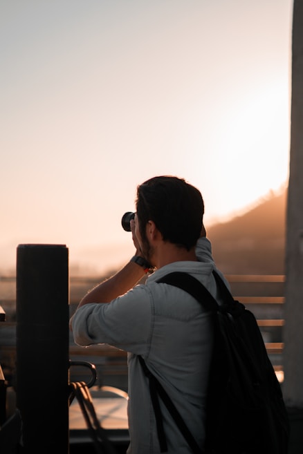 A person with a backpack is taking a photo with a camera against a sunset backdrop. The silhouette is partially illuminated by the warm glow of the setting sun, creating a serene and tranquil atmosphere.