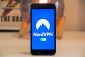 A smartphone displays the NordVPN logo in the center of its screen. The phone is positioned upright and placed on a light-colored surface with a blurred background that includes abstract elements.
