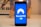 A smartphone displays the NordVPN logo in the center of its screen. The phone is positioned upright and placed on a light-colored surface with a blurred background that includes abstract elements.