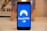 A smartphone displays the NordVPN logo in the center of its screen. The phone is positioned upright and placed on a light-colored surface with a blurred background that includes abstract elements.