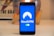 A smartphone displays the NordVPN logo in the center of its screen. The phone is positioned upright and placed on a light-colored surface with a blurred background that includes abstract elements.