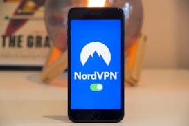 A smartphone displays the NordVPN logo in the center of its screen. The phone is positioned upright and placed on a light-colored surface with a blurred background that includes abstract elements.