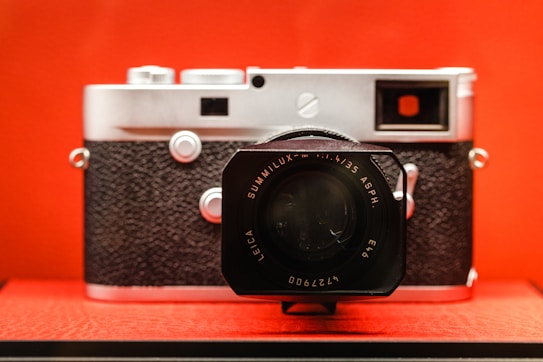 A vintage-style camera with a black textured grip and silver metallic body is prominently displayed against a vibrant red background. A large lens with a detailed front element features engravings.