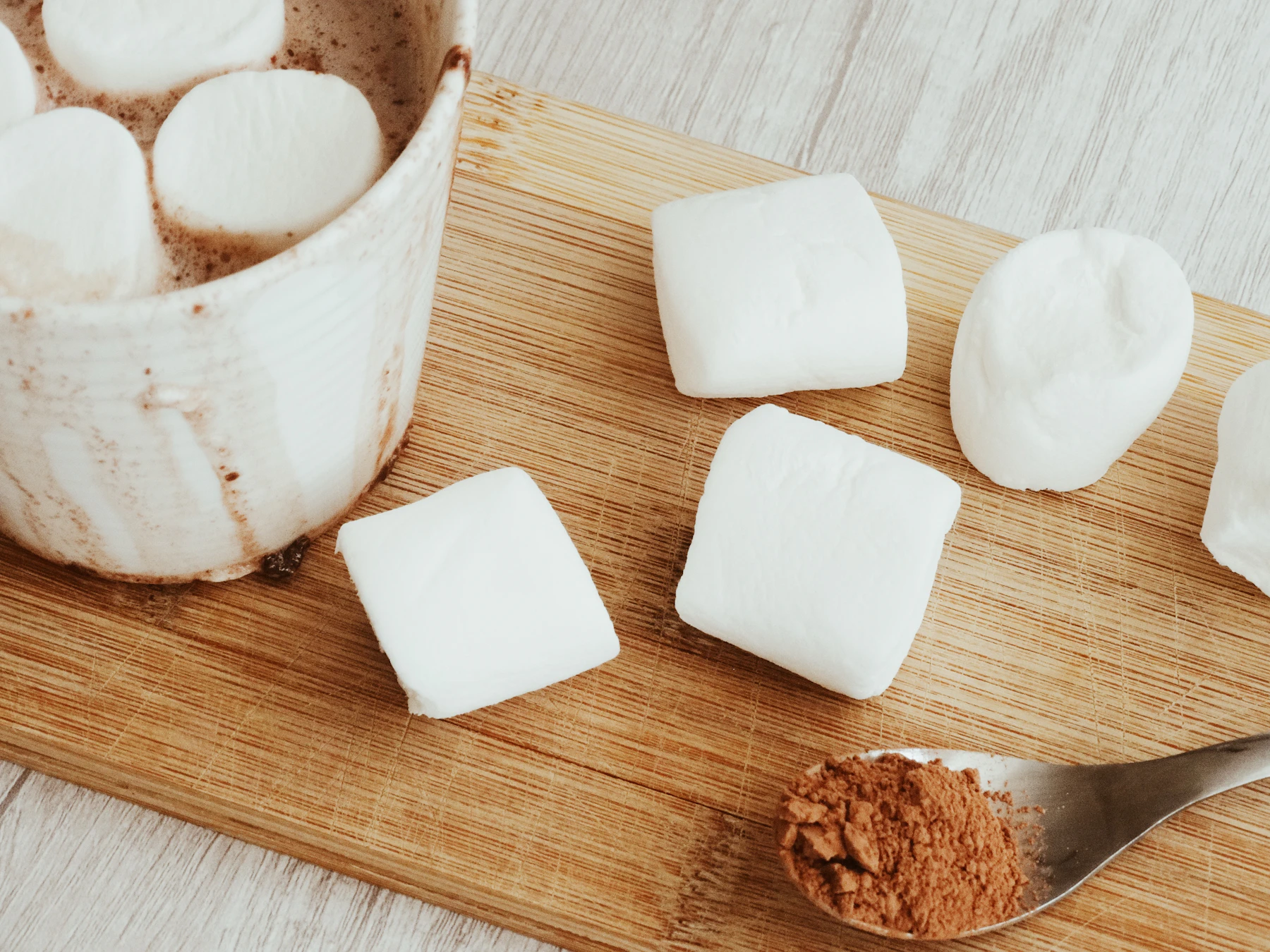 Square white marshmallows on a wooden board beside a mug of hot chocolate and a spoon of cocoa powder