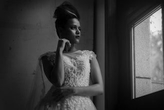 A person in an elegant, detailed dress gazes out a window with a thoughtful expression. The lighting is soft and creates a dramatic contrast, highlighting the intricate patterns of the dress and the person's hairstyle.