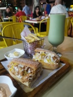 A delicious meal consisting of a sandwich filled with fried chicken and melted cheese is placed on a wooden tray. Beside the sandwich is a small basket of crispy french fries and a green smoothie in a tall glass. The setting appears to be a casual dining environment with yellow chairs and tables, and people in the background conversing.