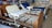 A room containing several hospital beds, some with attached bedside tables. The beds are equipped with adjustable rails and various types of mattresses. The room has posters and shelves lining the walls, filled with various medical and bedding supplies.