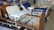 A room containing several hospital beds, some with attached bedside tables. The beds are equipped with adjustable rails and various types of mattresses. The room has posters and shelves lining the walls, filled with various medical and bedding supplies.
