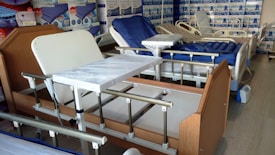 A room containing several hospital beds, some with attached bedside tables. The beds are equipped with adjustable rails and various types of mattresses. The room has posters and shelves lining the walls, filled with various medical and bedding supplies.