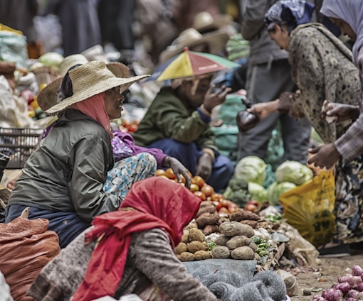 A bustling outdoor market scene with several people engaged in selling and buying fresh vegetables. Vendors sit on the ground surrounded by produce such as tomatoes, cabbages, potatoes, and green beans. Many of the individuals are dressed in layered clothing, and one person wears a multicolored umbrella hat.