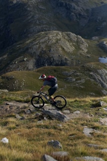A mountain biker is riding on a rocky and grassy trail, surrounded by rugged mountainous terrain. The biker wears a helmet and a backpack, navigating through a scenic landscape with a mix of green and brown tones.