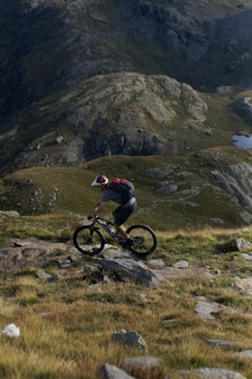A mountain biker is riding on a rocky and grassy trail, surrounded by rugged mountainous terrain. The biker wears a helmet and a backpack, navigating through a scenic landscape with a mix of green and brown tones.