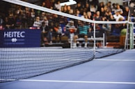 A tennis court is captured with focus on the net, stretching diagonally across the image. Spectators are in the background, seated and slightly out of focus. The court surface appears blue with visible white lines.