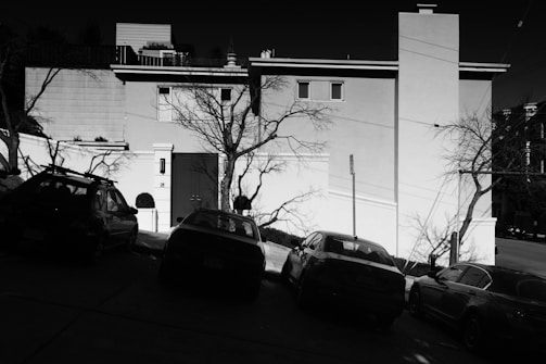 A black and white image depicting a residential area with several parked cars on a sloped street. The background features a large house with minimalistic architecture. Bare trees and their shadows create intricate patterns on the building's facade.