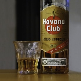 A bottle of Havana Club Añejo Especial rum stands next to a glass partially filled with the amber liquid. The setting is simple, with a flat surface beneath both the bottle and the glass, in an intimate low-light ambiance.