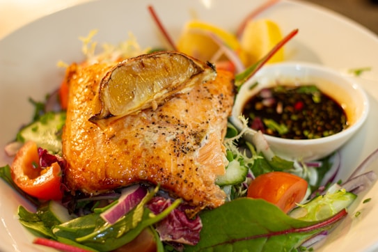 A plate featuring a piece of grilled salmon topped with a slice of lemon, resting on a colorful bed of fresh salad with tomatoes, cucumbers, and red onions. A small bowl of dipping sauce accompanies the dish, adding a touch of elegance.