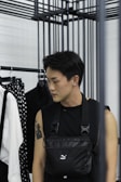 A person with short dark hair is standing beside a clothing rack. They are wearing a sleeveless black top and a black crossbody bag. The person has a tattoo on their left arm and an earring in their left ear. In the background, there is a white shirt with black suspenders and a polka-dot patterned item hanging on the rack.