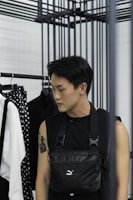 A person with short dark hair is standing beside a clothing rack. They are wearing a sleeveless black top and a black crossbody bag. The person has a tattoo on their left arm and an earring in their left ear. In the background, there is a white shirt with black suspenders and a polka-dot patterned item hanging on the rack.