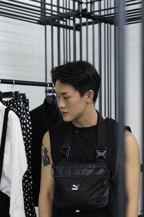 A person with short dark hair is standing beside a clothing rack. They are wearing a sleeveless black top and a black crossbody bag. The person has a tattoo on their left arm and an earring in their left ear. In the background, there is a white shirt with black suspenders and a polka-dot patterned item hanging on the rack.