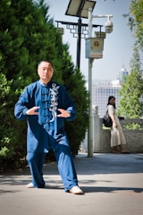 A person is performing Tai Chi in a park setting, wearing a blue traditional martial arts outfit. The surrounding area features lush greenery and a modern surveillance camera mounted on a pole. In the background, a woman in a white dress is leaning on a stone railing, possibly observing the exercise. Urban buildings are visible in the distant background under a clear sky.