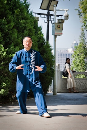 A person is performing Tai Chi in a park setting, wearing a blue traditional martial arts outfit. The surrounding area features lush greenery and a modern surveillance camera mounted on a pole. In the background, a woman in a white dress is leaning on a stone railing, possibly observing the exercise. Urban buildings are visible in the distant background under a clear sky.