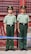 Two uniformed guards stand at attention in front of a weathered red door with metal bars. They are dressed in green military uniforms with matching hats featuring emblems.