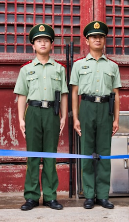 Two uniformed guards stand at attention in front of a weathered red door with metal bars. They are dressed in green military uniforms with matching hats featuring emblems.