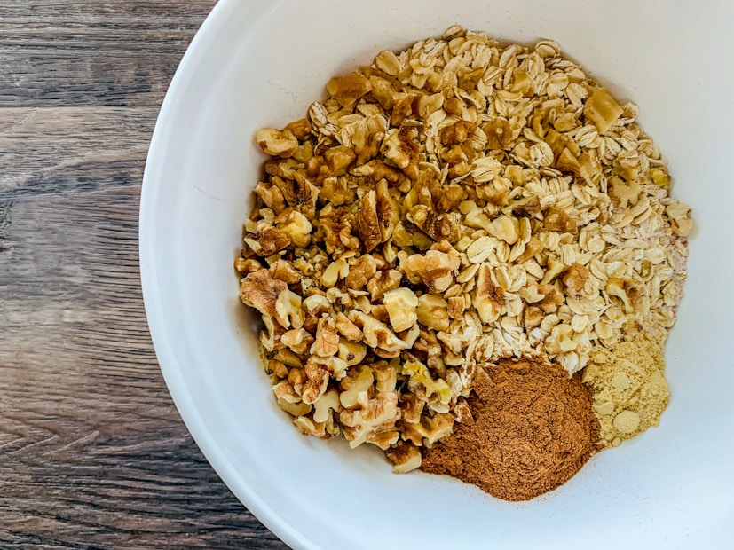 A white bowl contains a mixture of rolled oats, chopped walnuts, cinnamon, and ginger powder. The oats and walnuts are evenly distributed in the bowl, while the cinnamon and ginger powder are placed in small piles on one side. The bowl is set on a wooden surface with visible grain patterns.