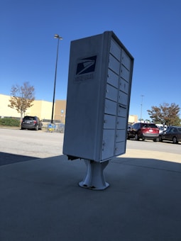 A cluster mailbox unit for the United States Postal Service is situated on a concrete surface under a clear blue sky. Several vehicles are parked in the background near some shopping carts, and there are a few trees with sparse foliage. Streetlights and a shopping cart return area are visible.