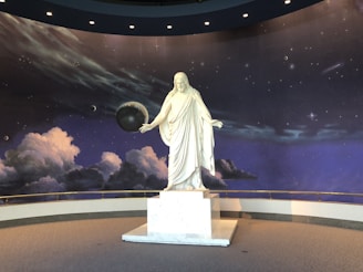 A white marble statue of a robed figure stands on a pedestal in front of a cosmic backdrop. The background features a starry sky with planets and swirling clouds, creating a celestial atmosphere.