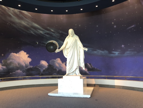 A white marble statue of a robed figure stands on a pedestal in front of a cosmic backdrop. The background features a starry sky with planets and swirling clouds, creating a celestial atmosphere.