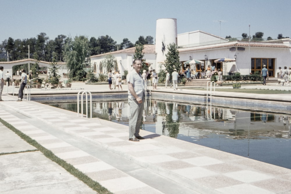 man in white shirt and gray pants standing beside body of water near white building during daytime