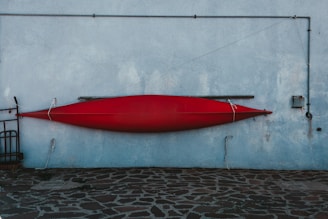 A red kayak is mounted on a textured light blue wall with metal brackets and ropes. The wall has metal pipes running along it, and a stone pavement with a rugged pattern lies beneath. The kayak is positioned horizontally, secured carefully.