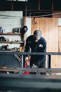 A person wearing a blue jumpsuit and protective gloves is engaged in a task involving a piece of equipment in a workshop. The setting has wooden and metal elements, shelves with various tools and items, and a helmet hanging on the wall.
