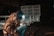 A person wearing protective gear and a camouflage jacket is welding a metal structure, with sparks flying in a dimly lit industrial setting. The metal frame looks like it could be part of a trailer or similar equipment.