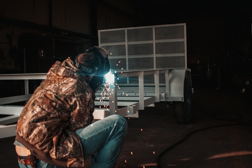 A person wearing protective gear and a camouflage jacket is welding a metal structure, with sparks flying in a dimly lit industrial setting. The metal frame looks like it could be part of a trailer or similar equipment.