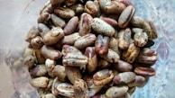 A close-up view of a pile of coffee beans with a raw and unroasted appearance. The beans have a mix of brown and dusty cream colors, with some surface imperfections visible. The background appears slightly blurred, emphasizing the texture and details of the beans.