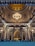 An ornate mosque interior featuring a large golden dome adorned with intricate designs and a central chandelier. The architecture includes arches and detailed wall patterns. A congregation of people is seated on a blue-carpeted floor, facing a decorative mihrab at the far end.