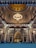An ornate mosque interior featuring a large golden dome adorned with intricate designs and a central chandelier. The architecture includes arches and detailed wall patterns. A congregation of people is seated on a blue-carpeted floor, facing a decorative mihrab at the far end.