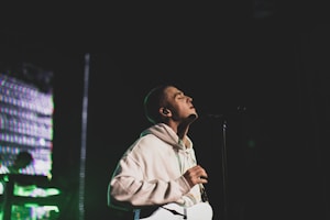 A person wearing a light-colored hoodie is passionately singing or speaking into a microphone on stage. The background features colorful lights and blurred elements, suggesting a performance atmosphere.