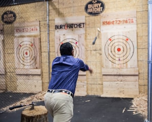 A man in a blue plaid shirt is engaged in axe throwing, aiming at a wooden target board with multiple concentric circles. Surrounding the area is a fenced enclosure, and an axe is embedded in a stump nearby.