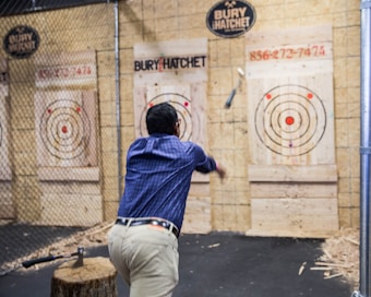 A man in a blue plaid shirt is engaged in axe throwing, aiming at a wooden target board with multiple concentric circles. Surrounding the area is a fenced enclosure, and an axe is embedded in a stump nearby.