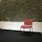 A single red upholstered chair with black metal legs is placed against a textured stone wall. The lower half of the wall is painted white, while the upper section showcases an exposed stone design. The floor appears dark, possibly a black carpet or flooring.
