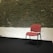 A single red upholstered chair with black metal legs is placed against a textured stone wall. The lower half of the wall is painted white, while the upper section showcases an exposed stone design. The floor appears dark, possibly a black carpet or flooring.
