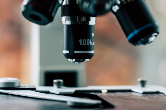 A close-up view of a microscope lens focusing on the stage. The microscope has an objective labeled with '100/1.25'. The background appears blurred, with warm hues that suggest a laboratory setting.