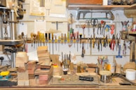 A cluttered workshop with a variety of tools hanging on a wall, including screwdrivers, pliers, and saws. There is a workbench in the foreground covered with wooden blocks, sandpaper, and various small tools and materials. The overall setting is that of a woodworking or crafting environment, with a drill press on the left and numerous objects organized and scattered throughout the space.
